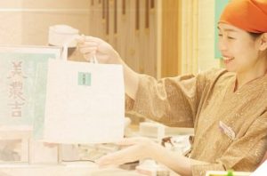 百貨店の食品売場で販売員が商品を丁寧に手渡す接客シーン。上質な和の雰囲気の中で行われる店頭販売商品紹介のイメージ