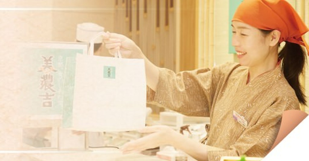 百貨店の食品売場で販売員が商品を丁寧に手渡す接客シーン。上質な和の雰囲気の中で行われる店頭販売商品紹介のイメージ